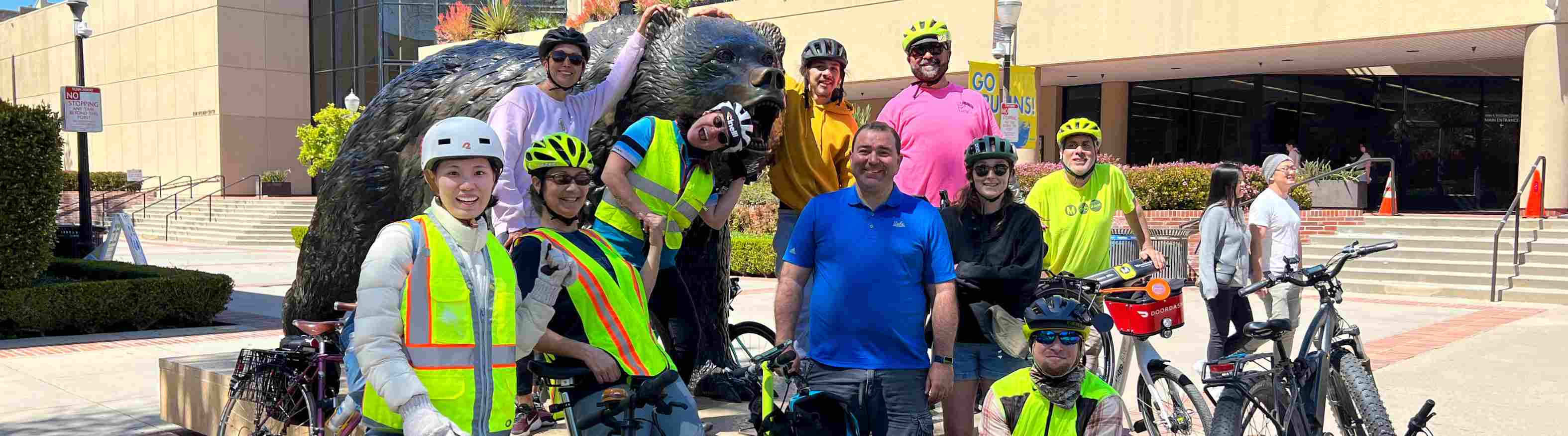 Happy riders with and without bikes smile in front of a sculpture of a bear. 