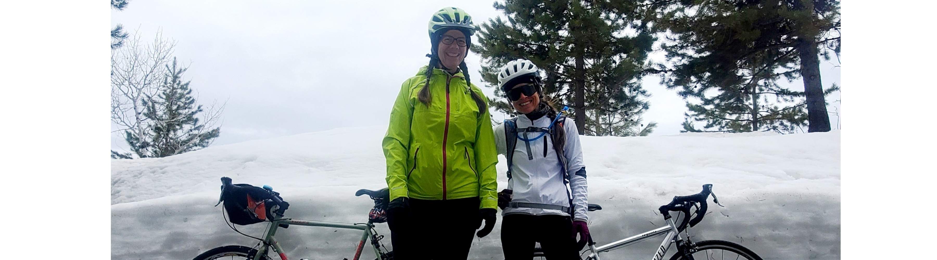 Two women standing next to their bikes in front of a big pile of snow